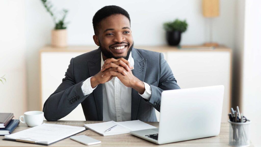 A man in a suit sits at a desk with a laptop, notepad, and cup, smiling as he considers B2B printing services Spokane. The office background includes plants and shelves.