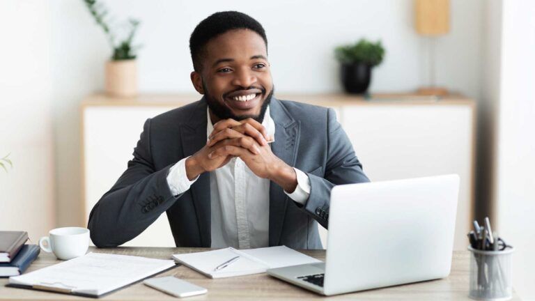 A man in a suit sits at a desk with a laptop, notepad, and cup, smiling as he considers B2B printing services Spokane. The office background includes plants and shelves.