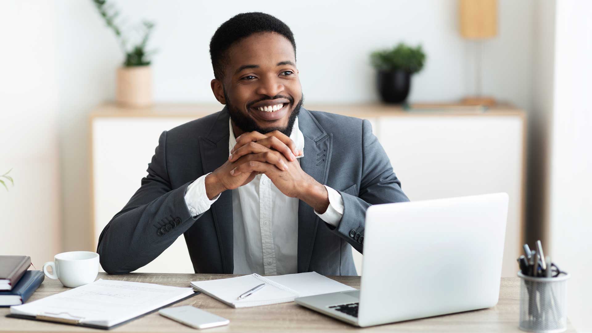 A man in a suit sits at a desk with a laptop, notepad, and cup, smiling as he considers B2B printing services Spokane. The office background includes plants and shelves.
