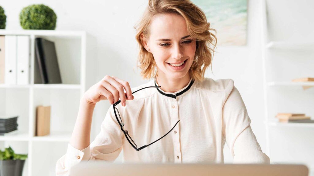 A woman with short blonde hair, wearing a light blouse, sits at a desk holding eyeglasses and smiling at a laptop screen—perhaps reviewing B2B printing services Spokane offers. Shelves with books and plants are visible in the background.