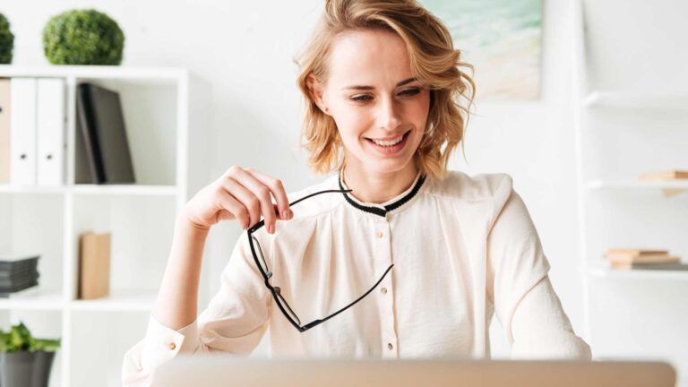 A woman with short blonde hair, wearing a light blouse, sits at a desk holding eyeglasses and smiling at a laptop screen—perhaps reviewing B2B printing services Spokane offers. Shelves with books and plants are visible in the background.