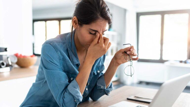 A woman sits at a table with a laptop, appearing stressed as she thinks about managing bulk newsletter printing for her business. Sunlight streams through large windows in the background, highlighting her need for B2B printing services Spokane.