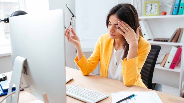 A woman in a yellow blazer sits at a desk in front of a computer, holding her glasses and rubbing her forehead, appearing stressed—perhaps overwhelmed by bulk newsletter printing tasks. Papers and office supplies are scattered around her.