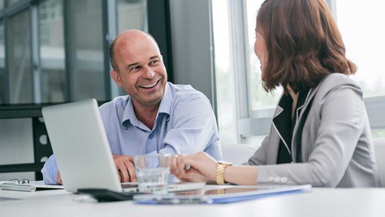 A man and a woman in business attire sit at a desk with a laptop and documents, discussing B2B printing services Spokane, smiling as they collaborate in a brightly lit office environment.