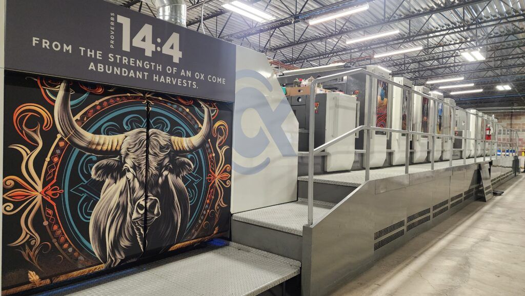 A large industrial printer for bulk newsletter printing stands in a warehouse, featuring a mural of an ox and patterns with the text, "Proverbs 14:4 From the strength of an ox come abundant harvests.